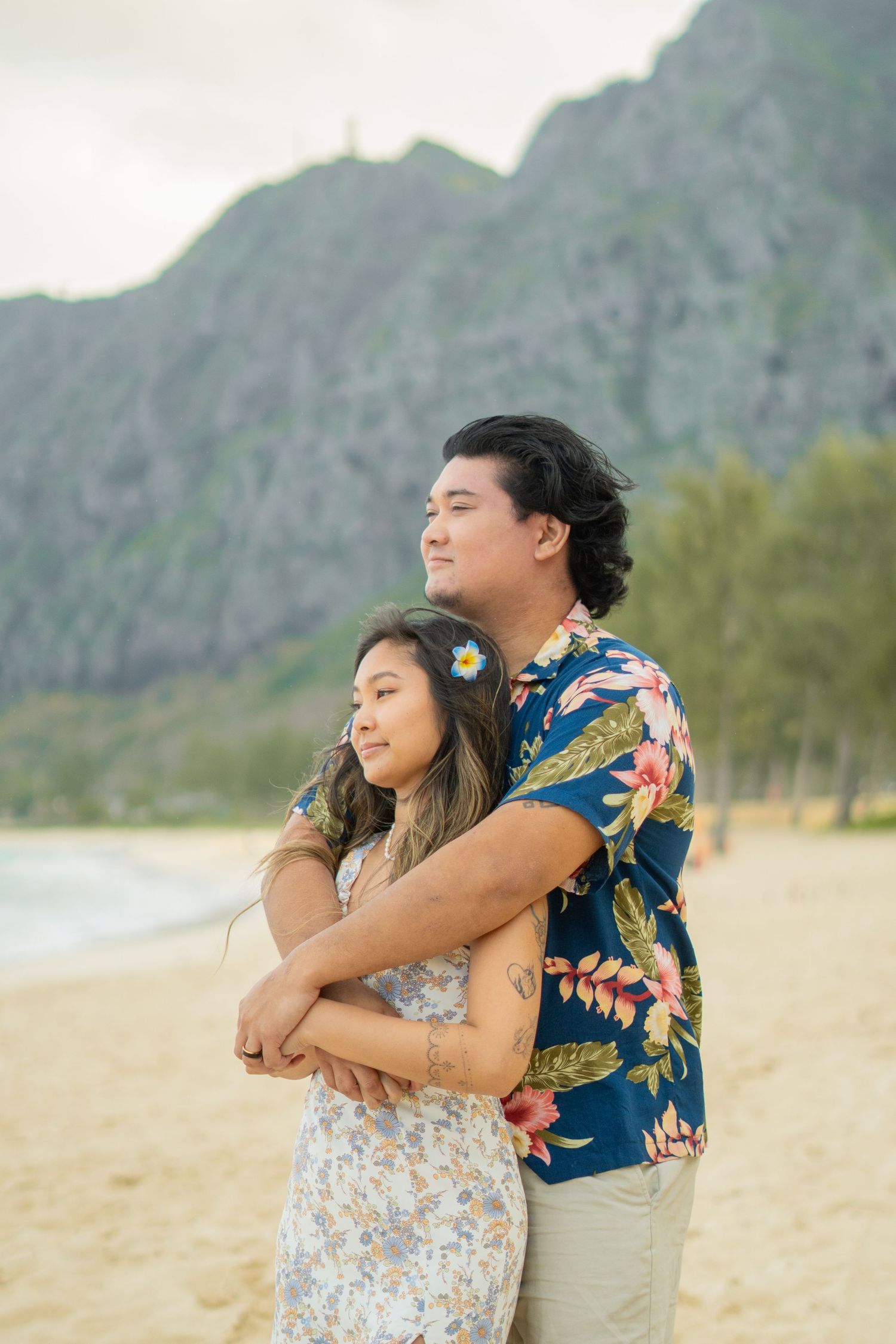 Couple in Hawaiian outfits embracing on sandy beach with lush green mountain backdrop in Hawaii.