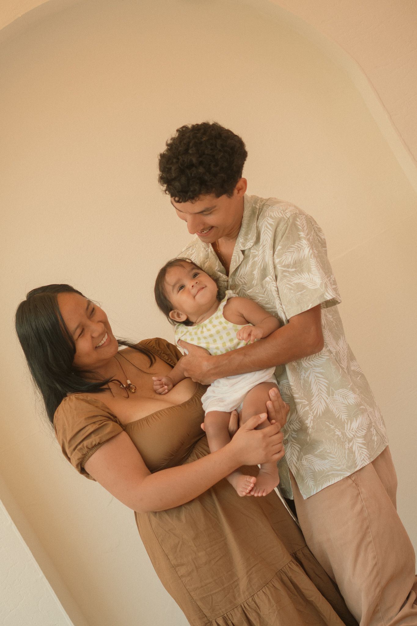 Happy young family with mother, father, and baby posing together in a warm-toned studio portrait.