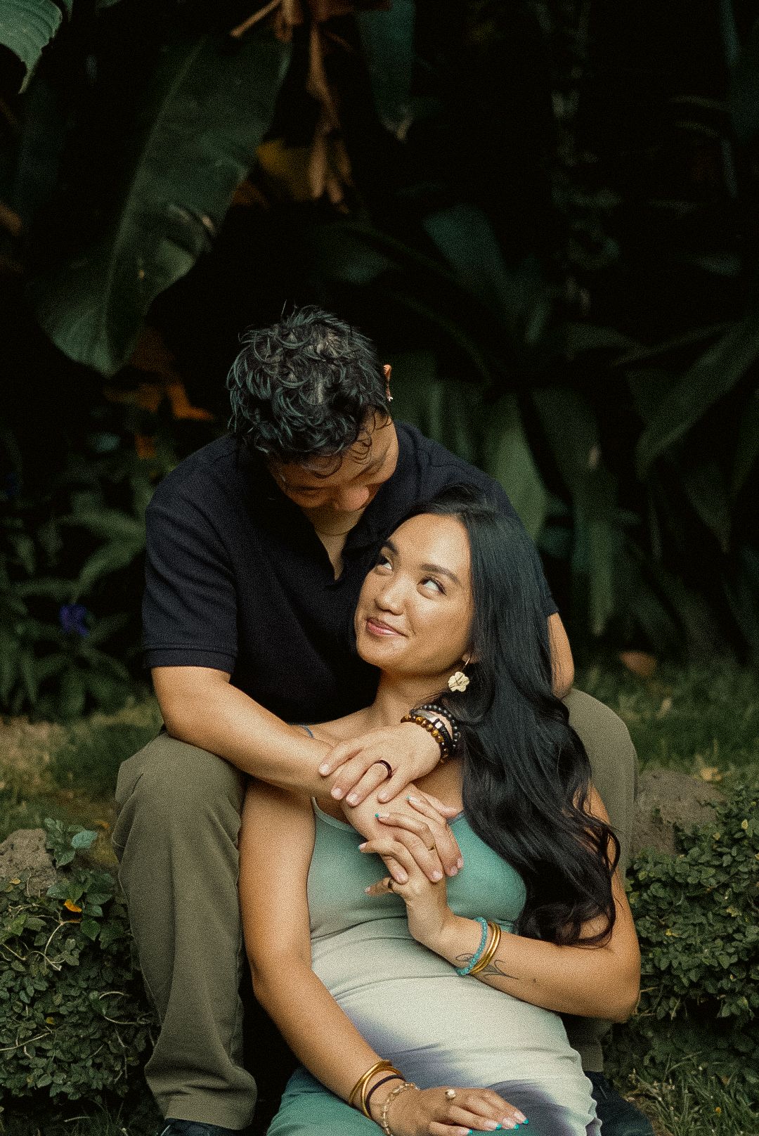 Couple shares a tender embrace while sitting among lush tropical greenery outdoors.