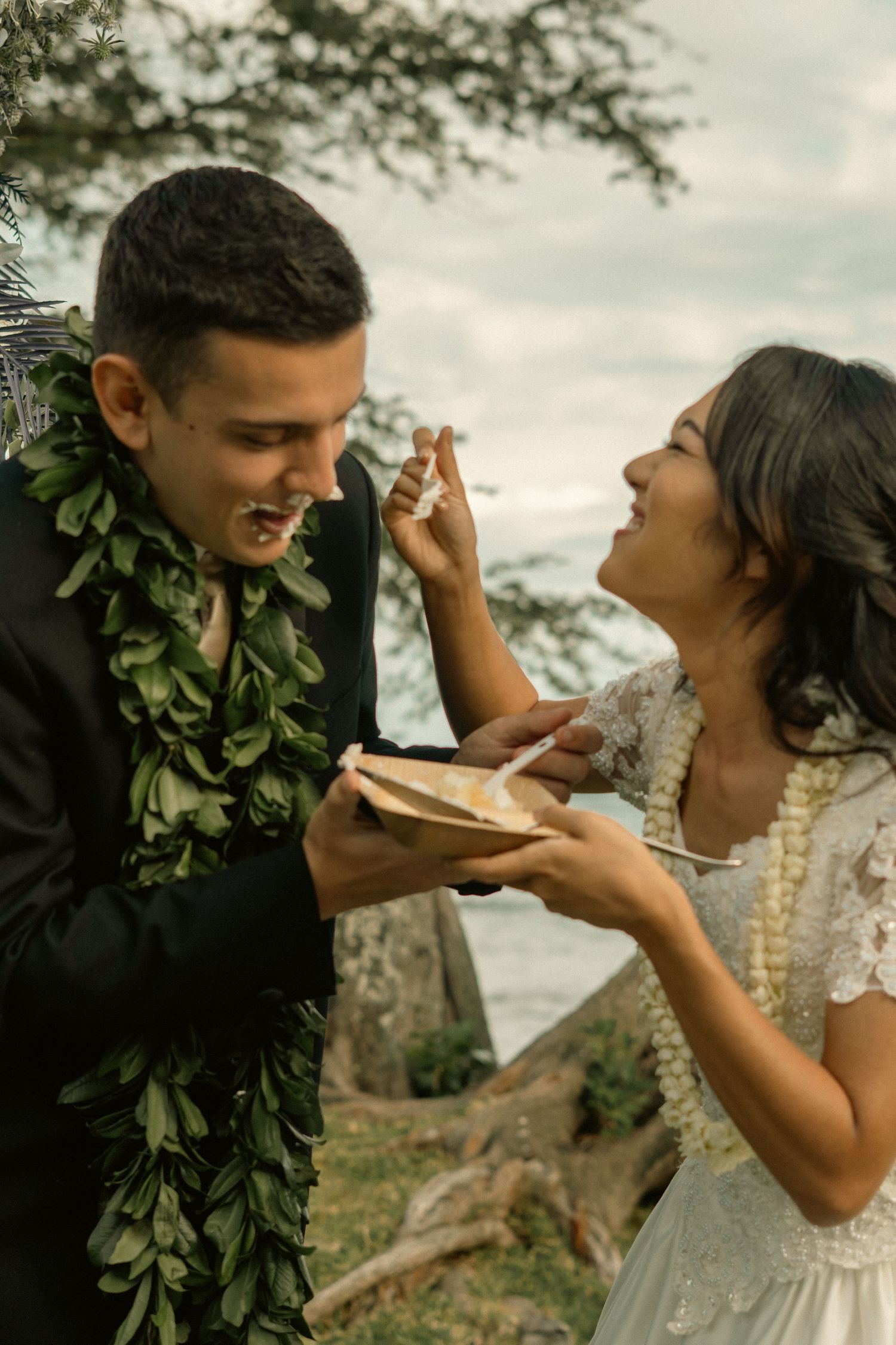 Couple in Hawaiian leis sharing wedding cake outdoors near ocean.