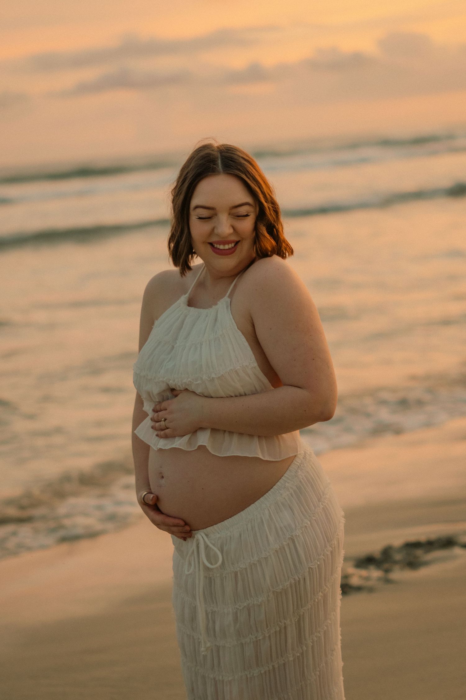 Pregnant woman in white crop top and skirt cradling her baby bump on a beach at golden sunset.