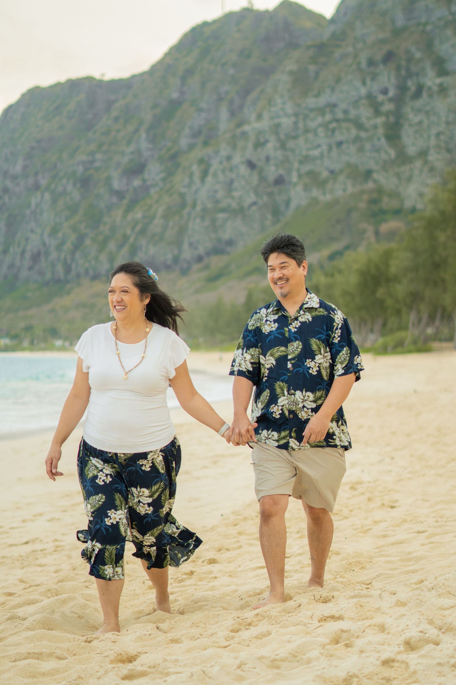 Smiling couple in Hawaiian attire walking hand in hand on sunny beach with mountain scenery.