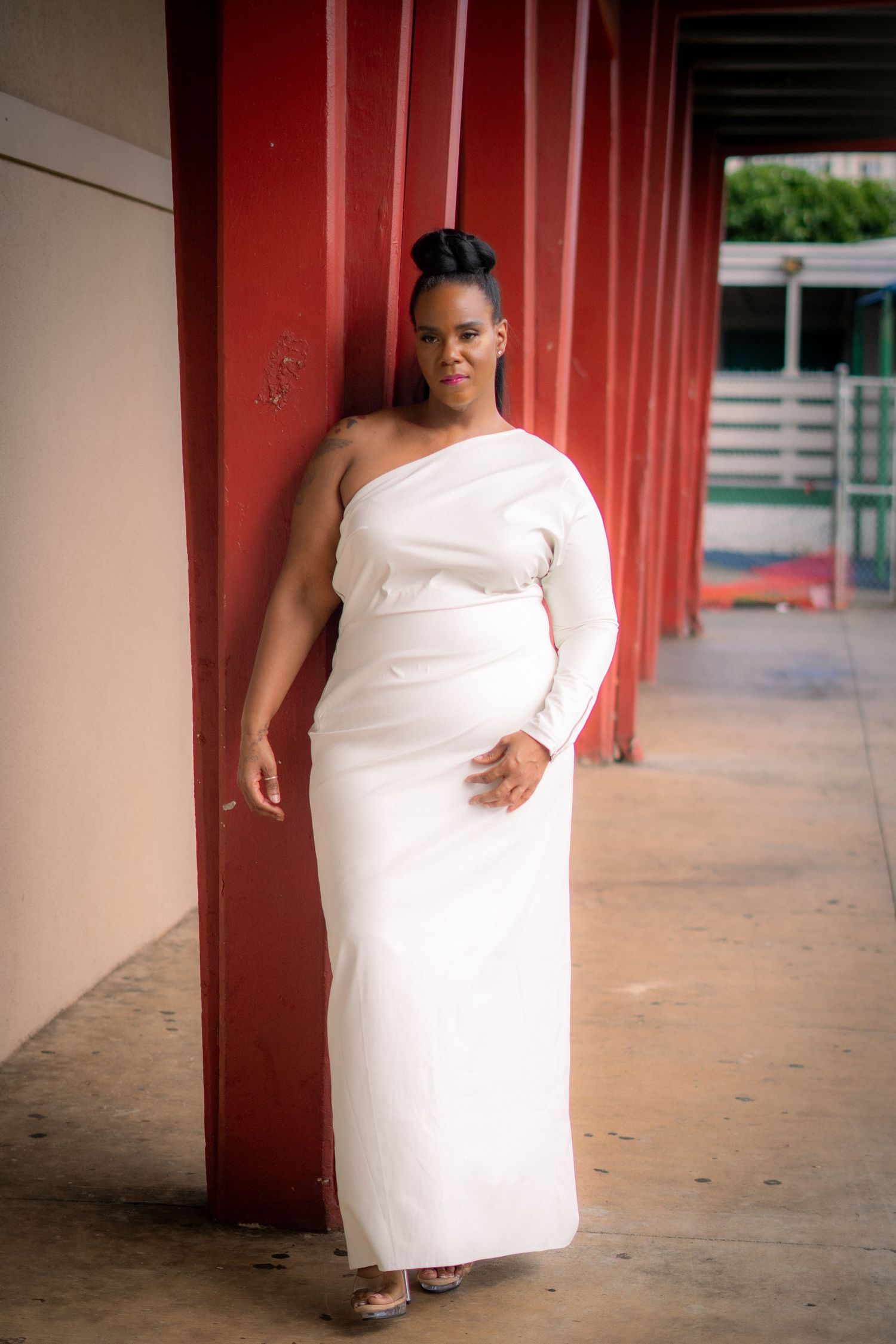 Elegant Black woman in white one-shoulder gown leaning against red column outdoors.