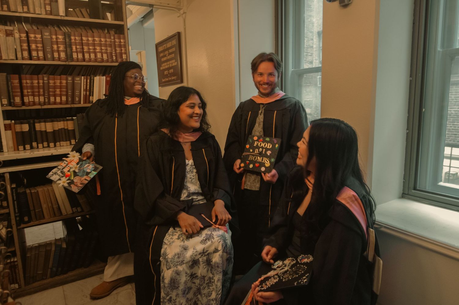 Four people in academic robes smiling and conversing in a library filled with old books.