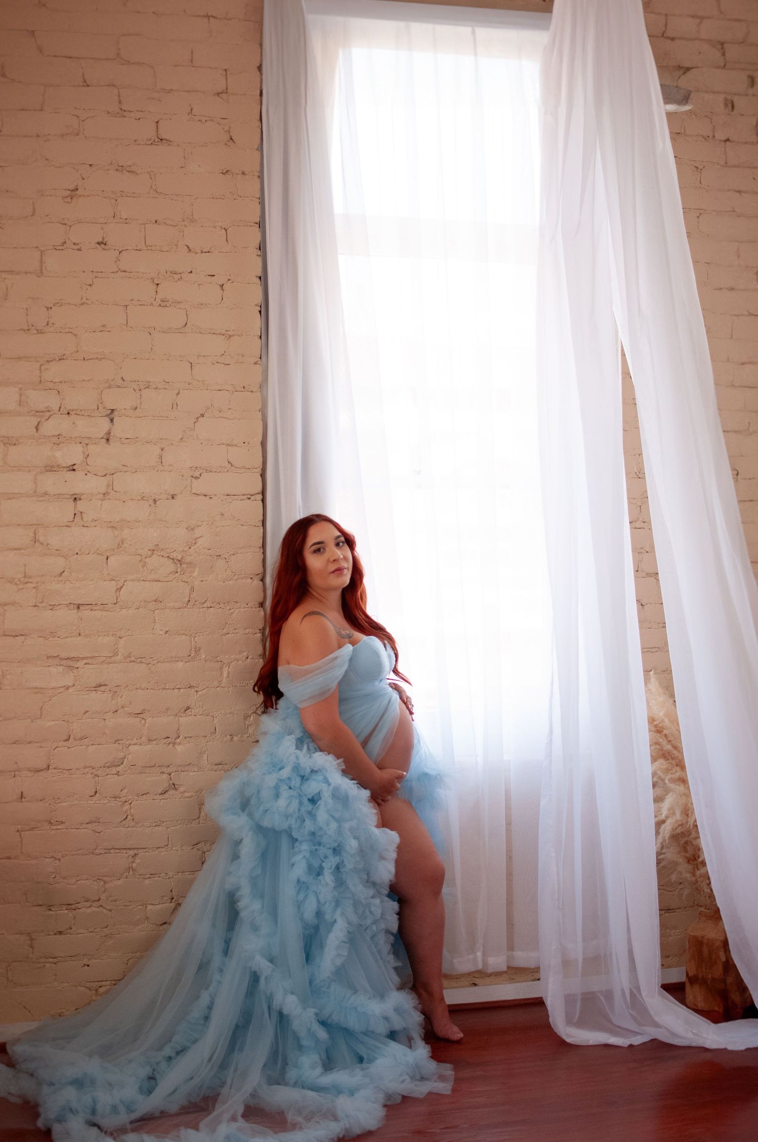 Pregnant woman in a flowing light blue tulle gown poses gracefully by a bright window with white curtains.