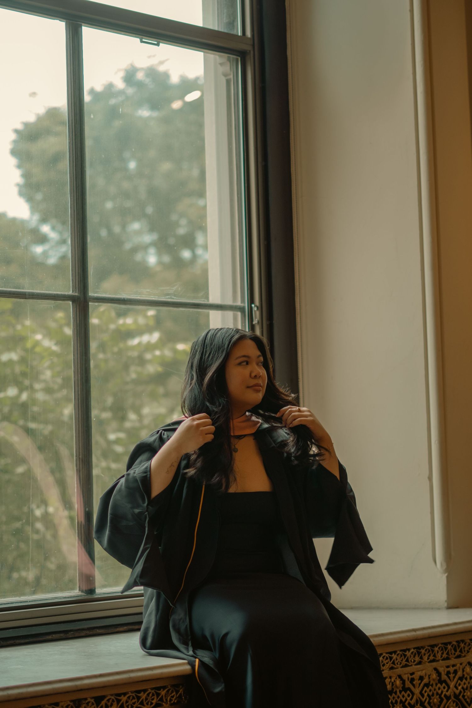 Woman in black graduation gown sits by a large window, gazing thoughtfully at lush green trees outside.
