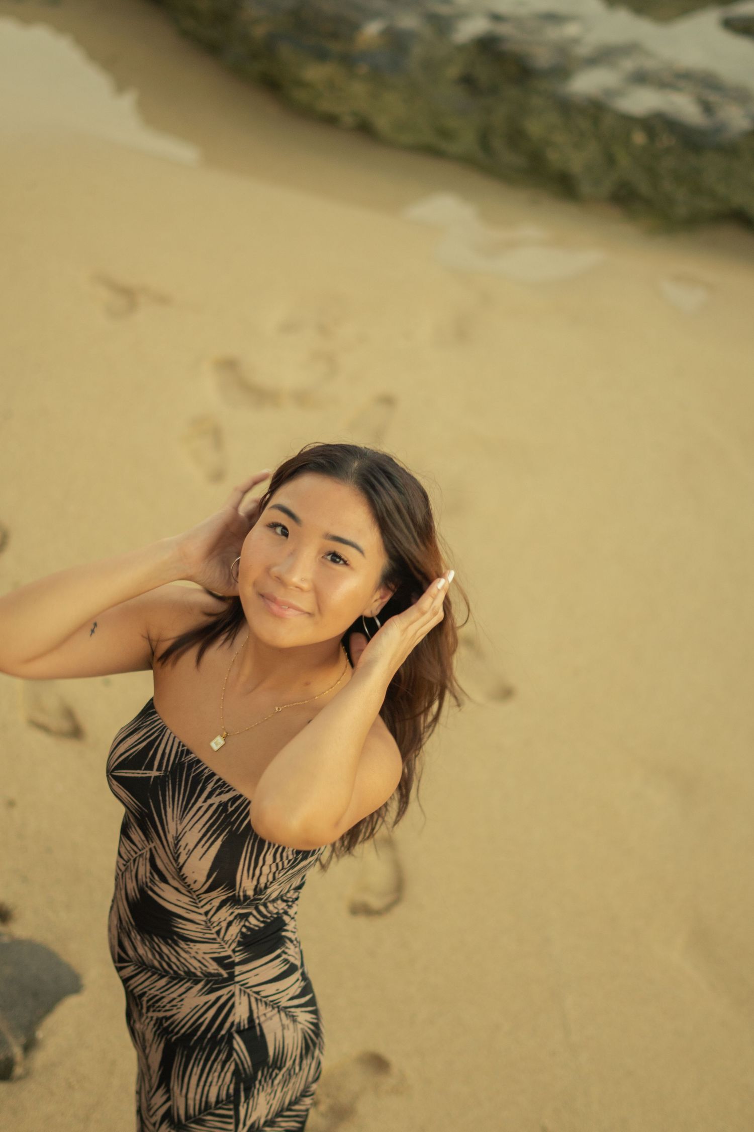 Asian woman in tropical print dress posing on sandy beach at golden hour with footprints behind her.