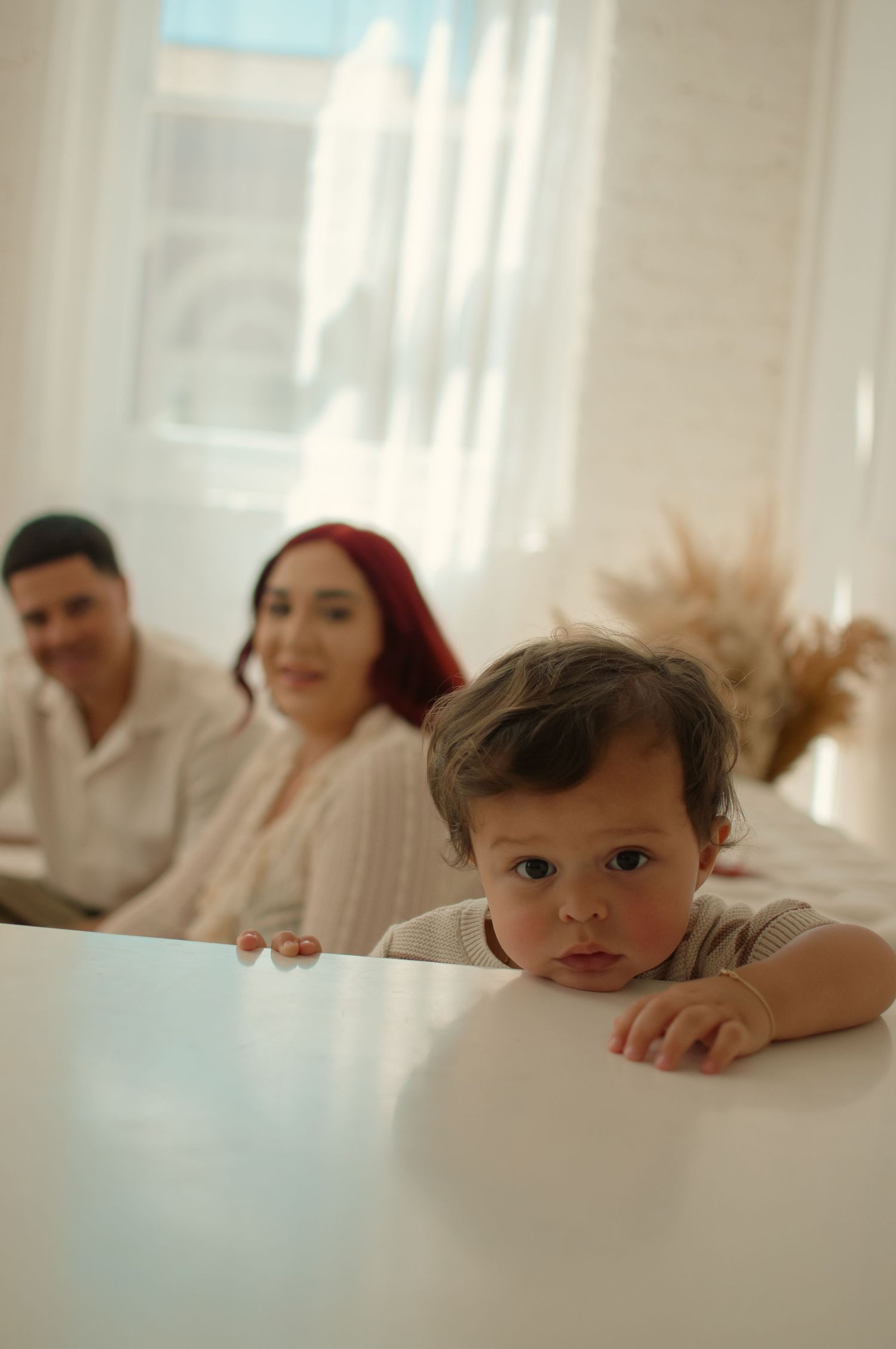 A curious toddler peers over a white table while his smiling parents sit blurred in the bright background.