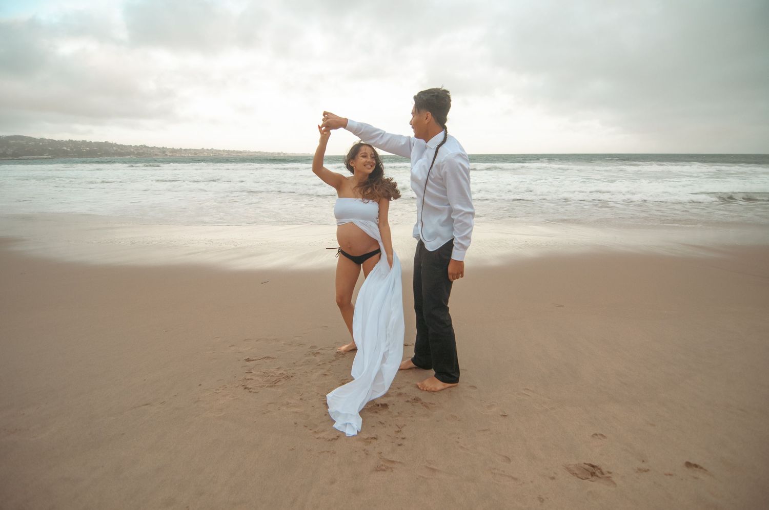 A couple dances on a beach with a flowing white fabric during a maternity photoshoot.