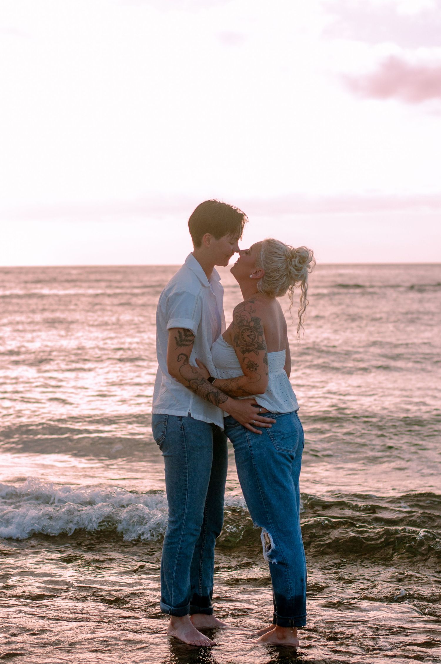 Tattooed couple shares romantic moment standing in ocean waves at sunset.