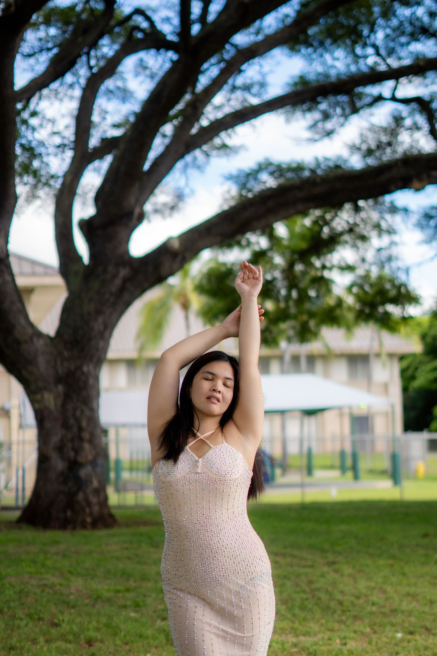 Young Asian woman in white dress stretching arms overhead under large tree outdoors.