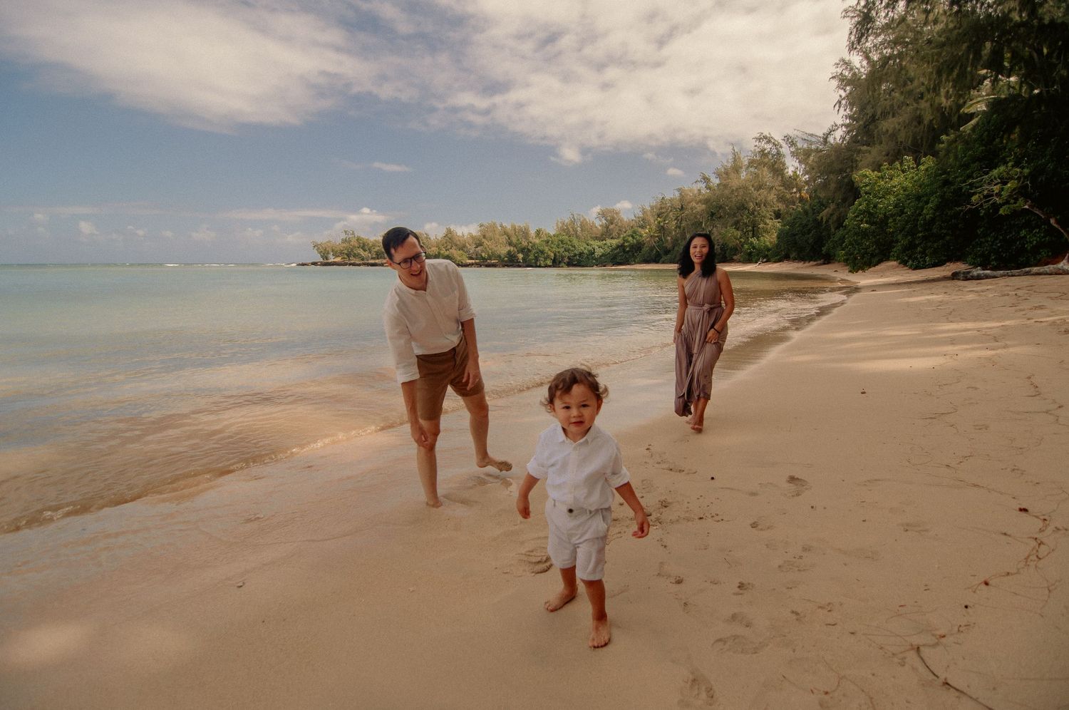 Family enjoying a sunny tropical beach walk with a toddler running on white sand near lush green trees.