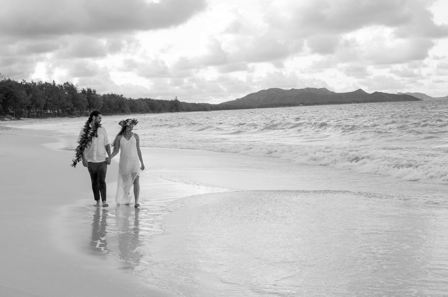 Two women walk hand-in-hand along a tropical beach shoreline in a black and white wedding photo.