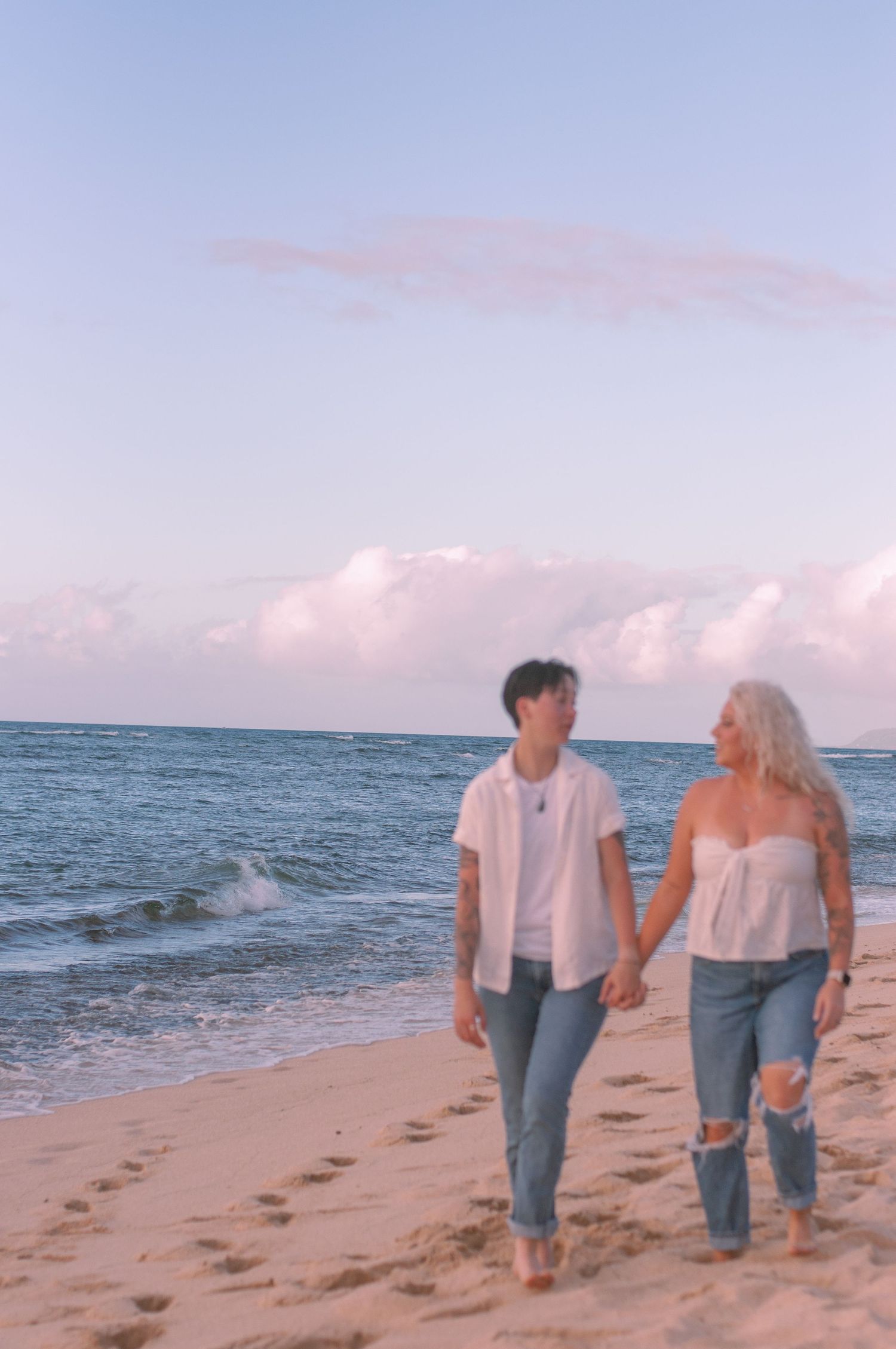 Couple walks hand in hand barefoot along sandy beach at pastel sunset.