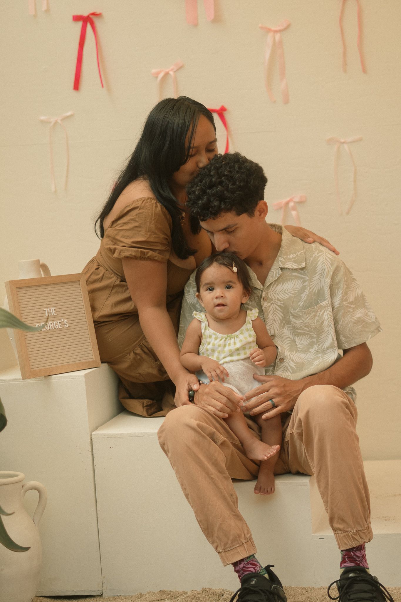 A young family poses for a photo, with a mother kissing father's cheek as they hold their baby girl.