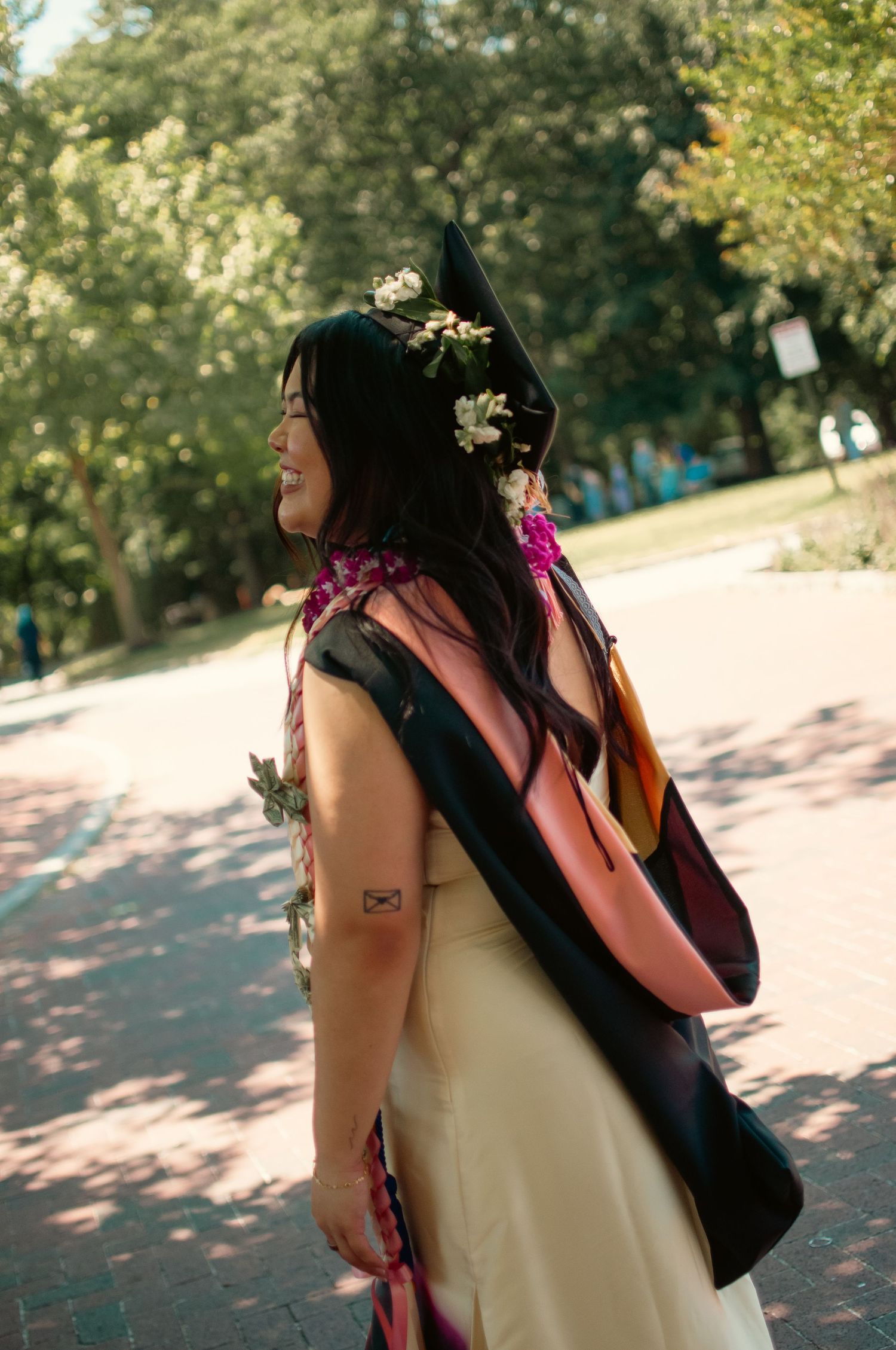 Smiling graduate in mortarboard hat adorned with flowers, wearing academic regalia over cream dress outdoors.
