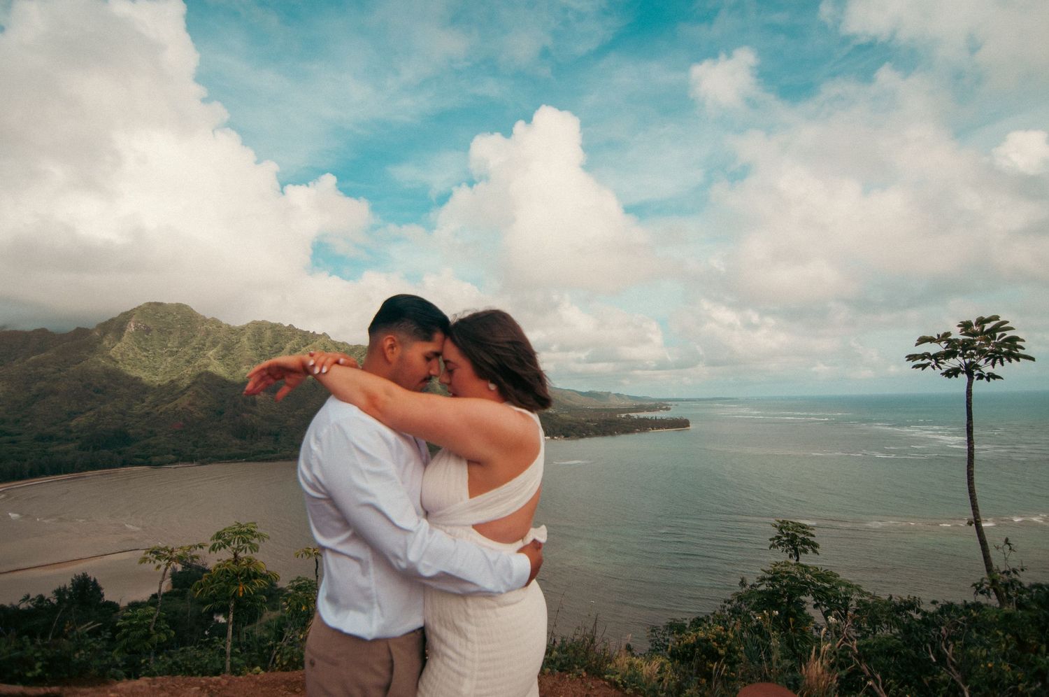 Couple embracing and kissing on a scenic overlook with lush mountains and ocean views under a cloudy sky.