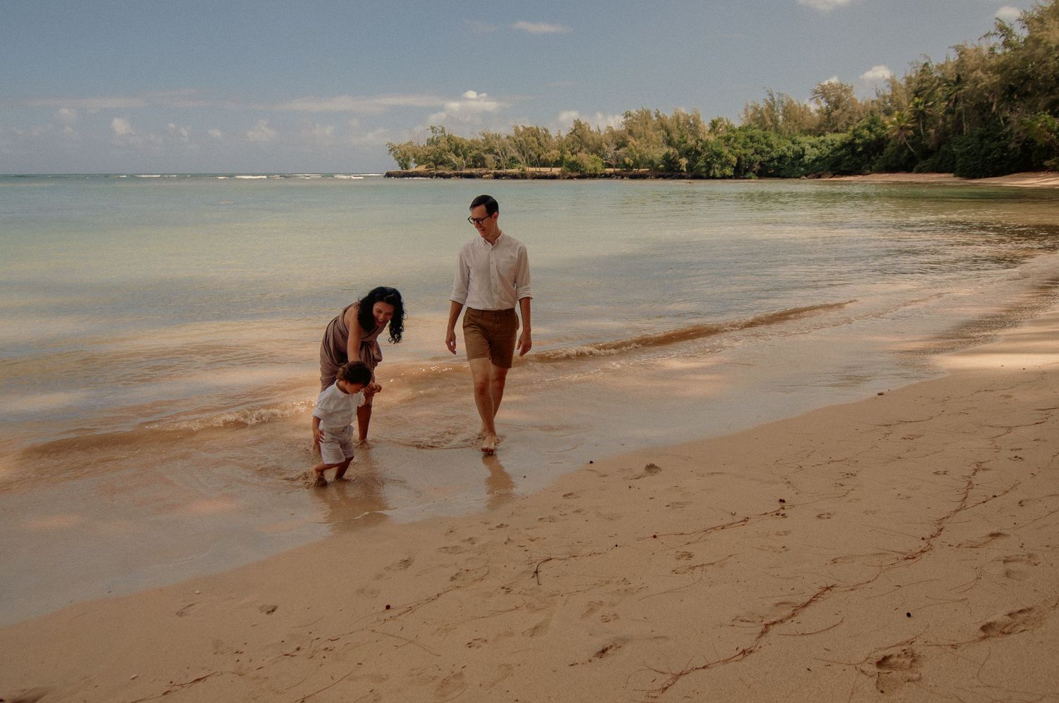 Couple walking along a tropical sandy beach shore with lush green trees and calm turquoise ocean waters.