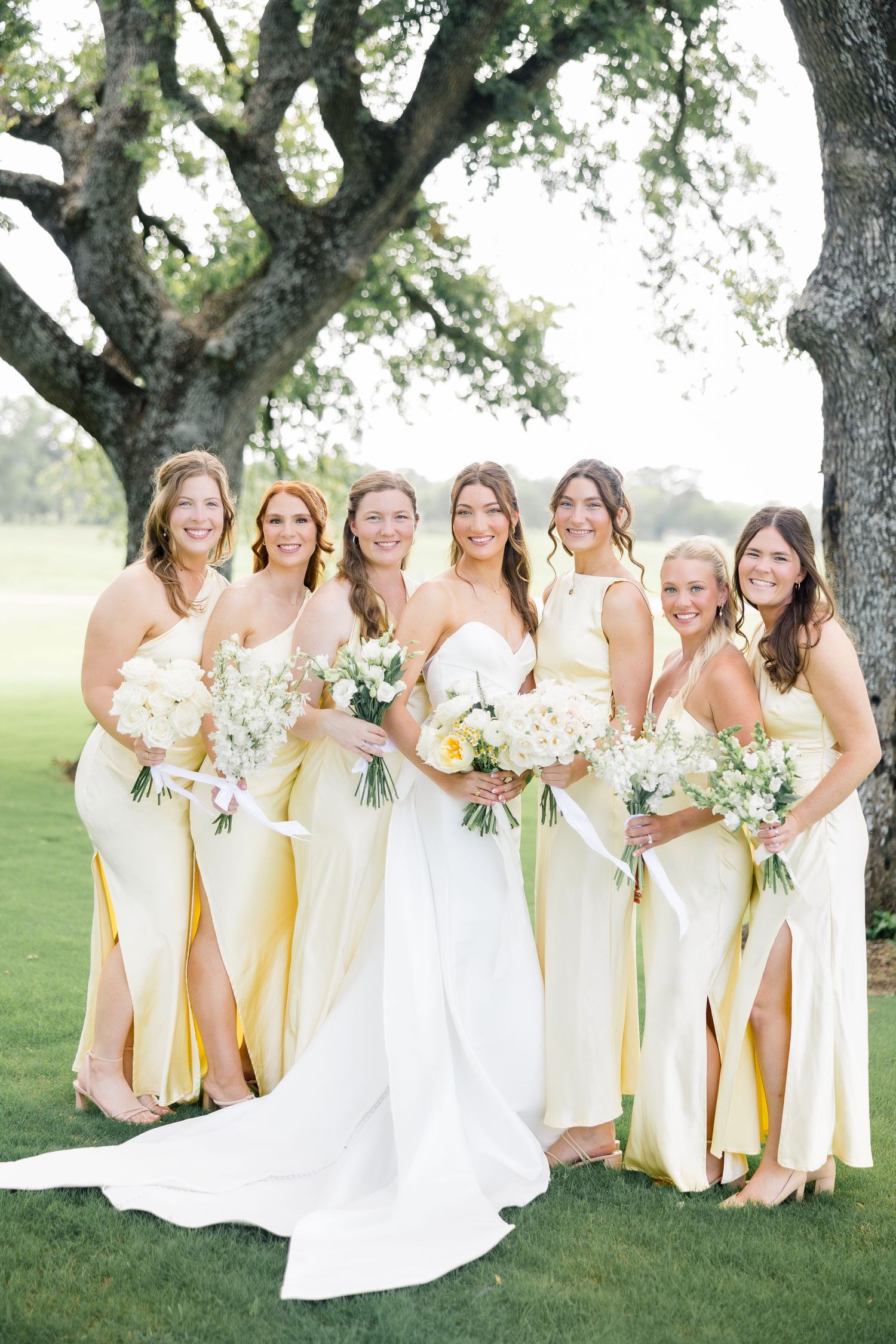 A group of bridesmaids in pale yellow dresses pose together with white floral bouquets on a grassy lawn. Chandelier Farms, Terrell TX.