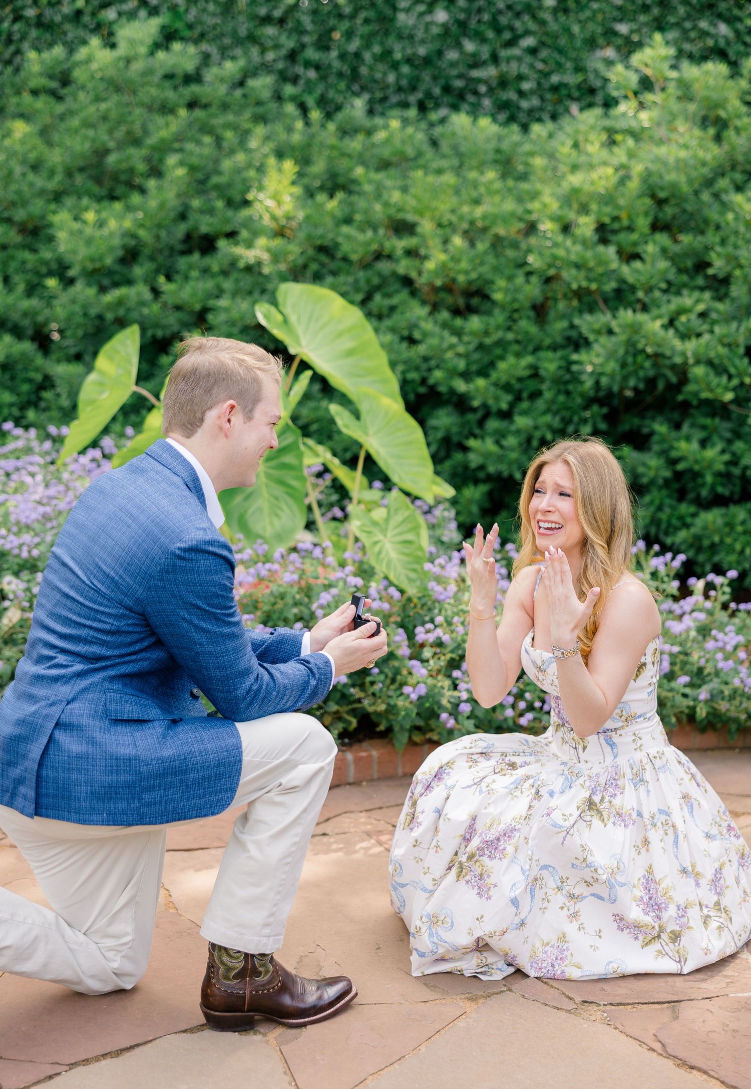 Man in blue blazer kneels to propose to woman in floral dress in a lush garden setting. Dallas arboretum engagement photos