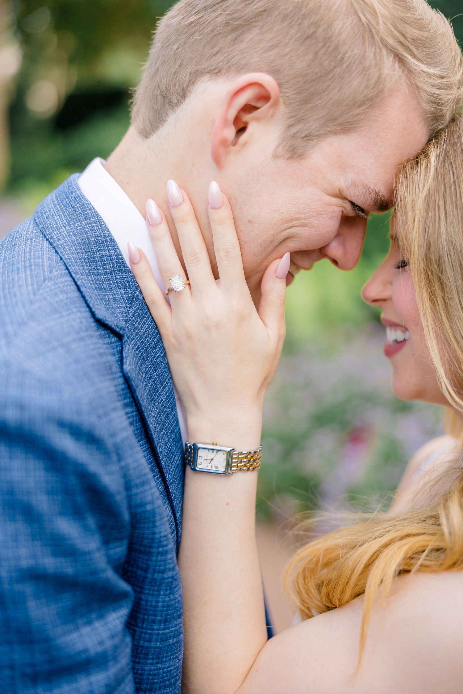 Couple sharing a romantic moment, woman showing engagement ring and gold watch, man in blue suit smiling at Dallas Arboretum
