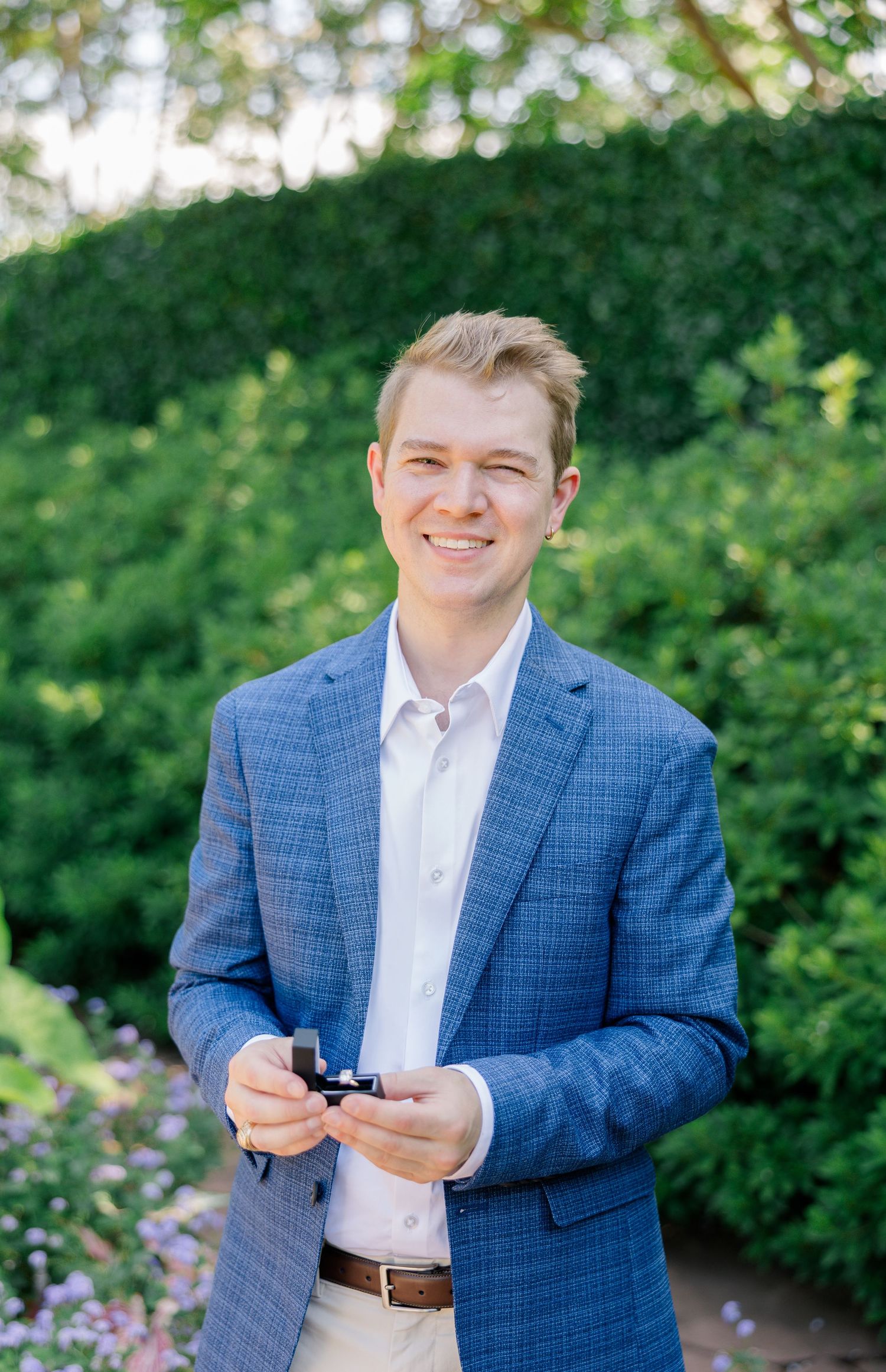 Young man in blue blazer smiling outdoors, holding a ring box before proposal at Dallas Arboretum.