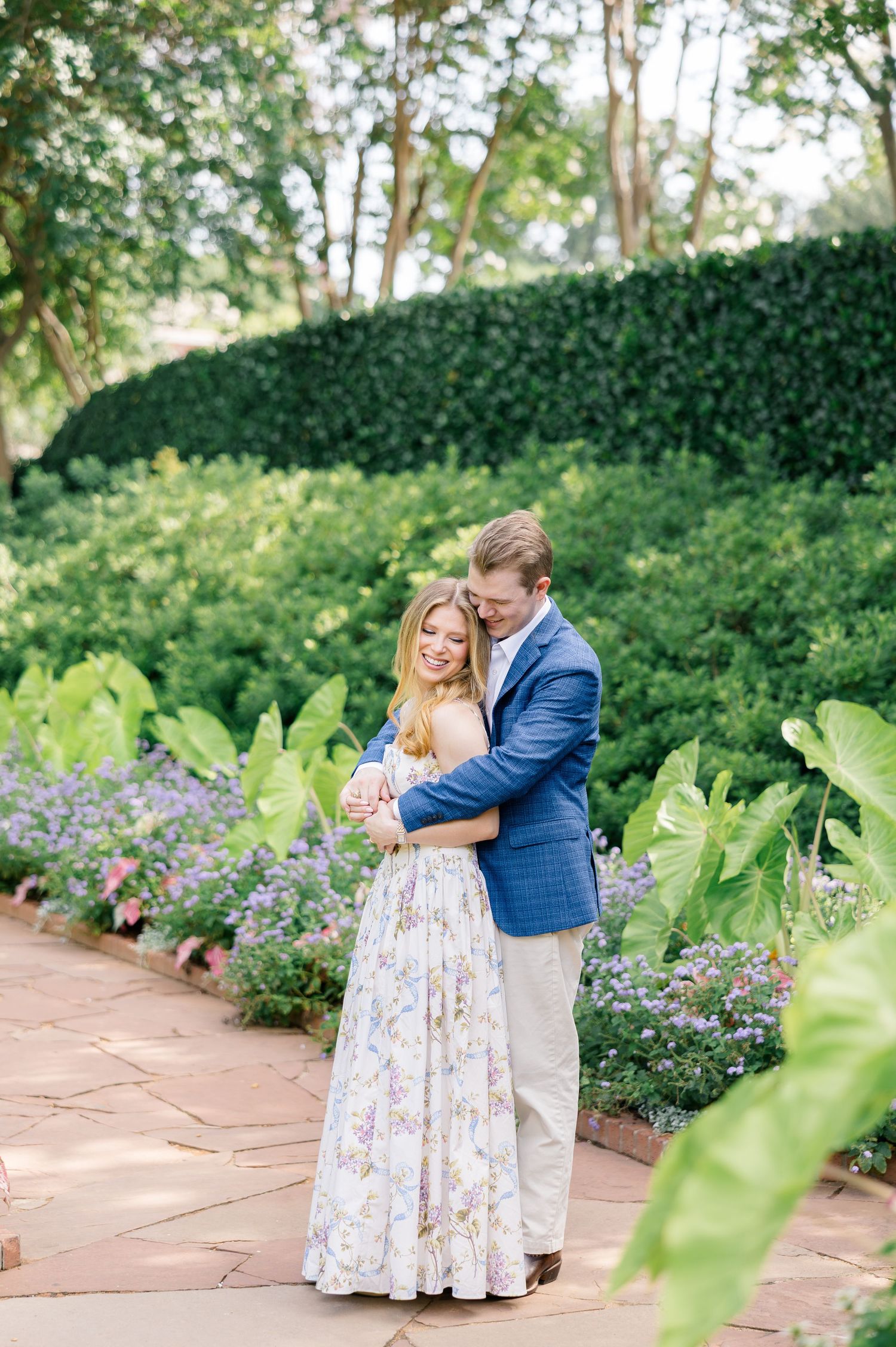 Couple embracing in a lush garden, woman in floral maxi dress and man in blue blazer on a brick pathway at Dallas Arboretum