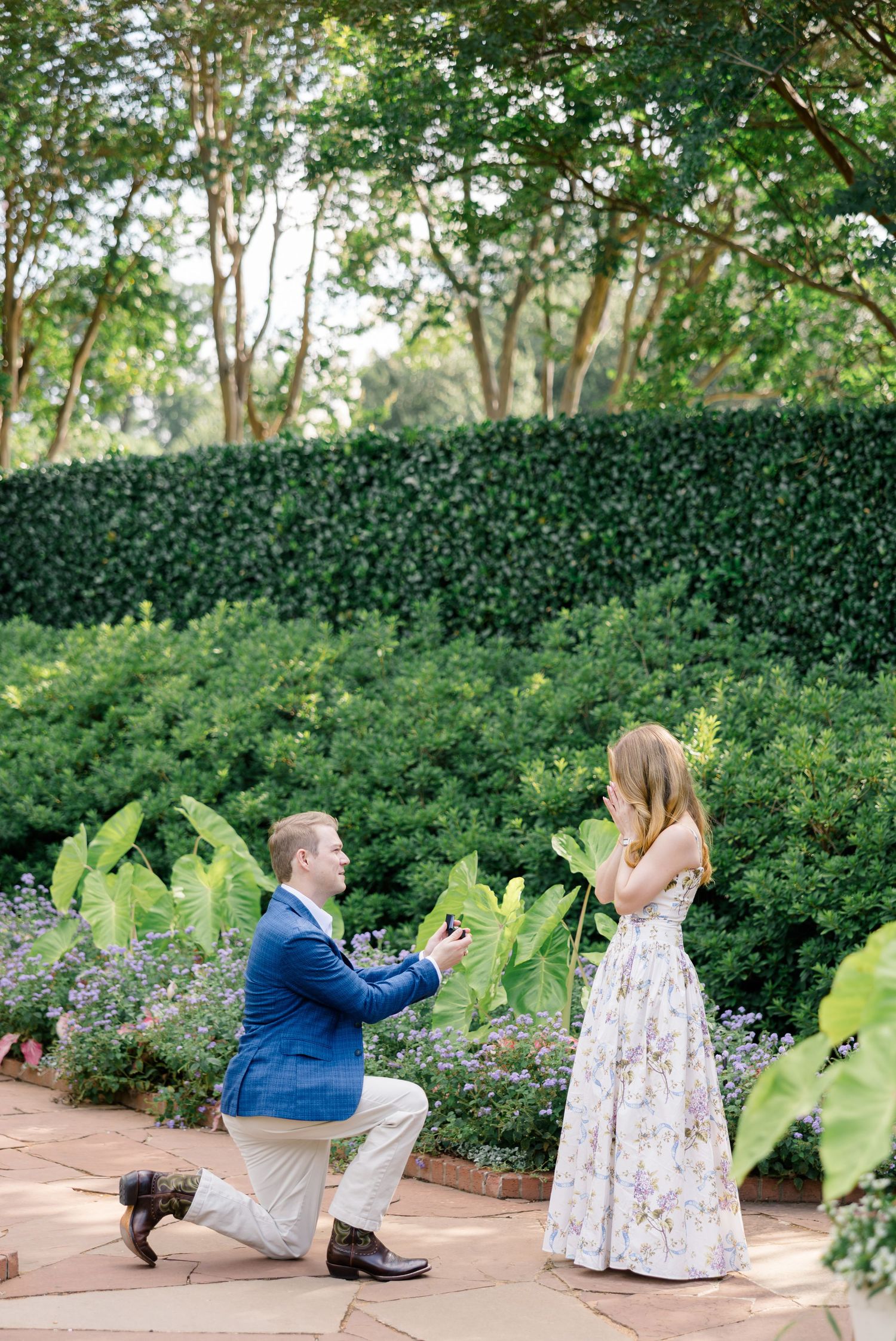 Man in blue suit proposes on one knee to woman in floral dress in a lush garden setting at Dallas Arboretum