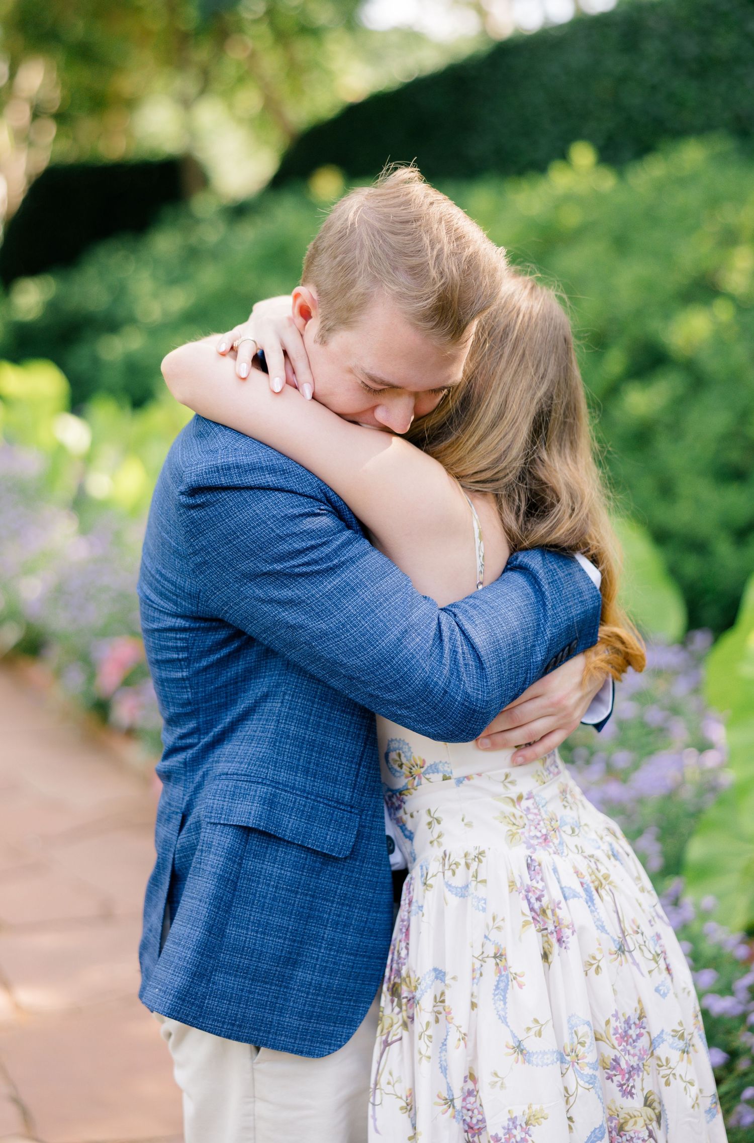 Couple embracing in a garden, man in blue blazer hugging woman in floral dress among lush greenery after romantic proposal in Dallas