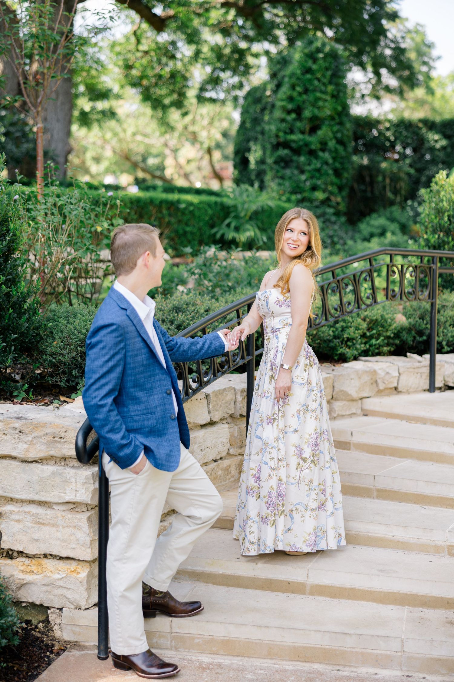 Couple holding hands on stone steps in a garden, man in blue blazer, woman in floral maxi dress at Dallas Arboretum Botanical Gardens.
