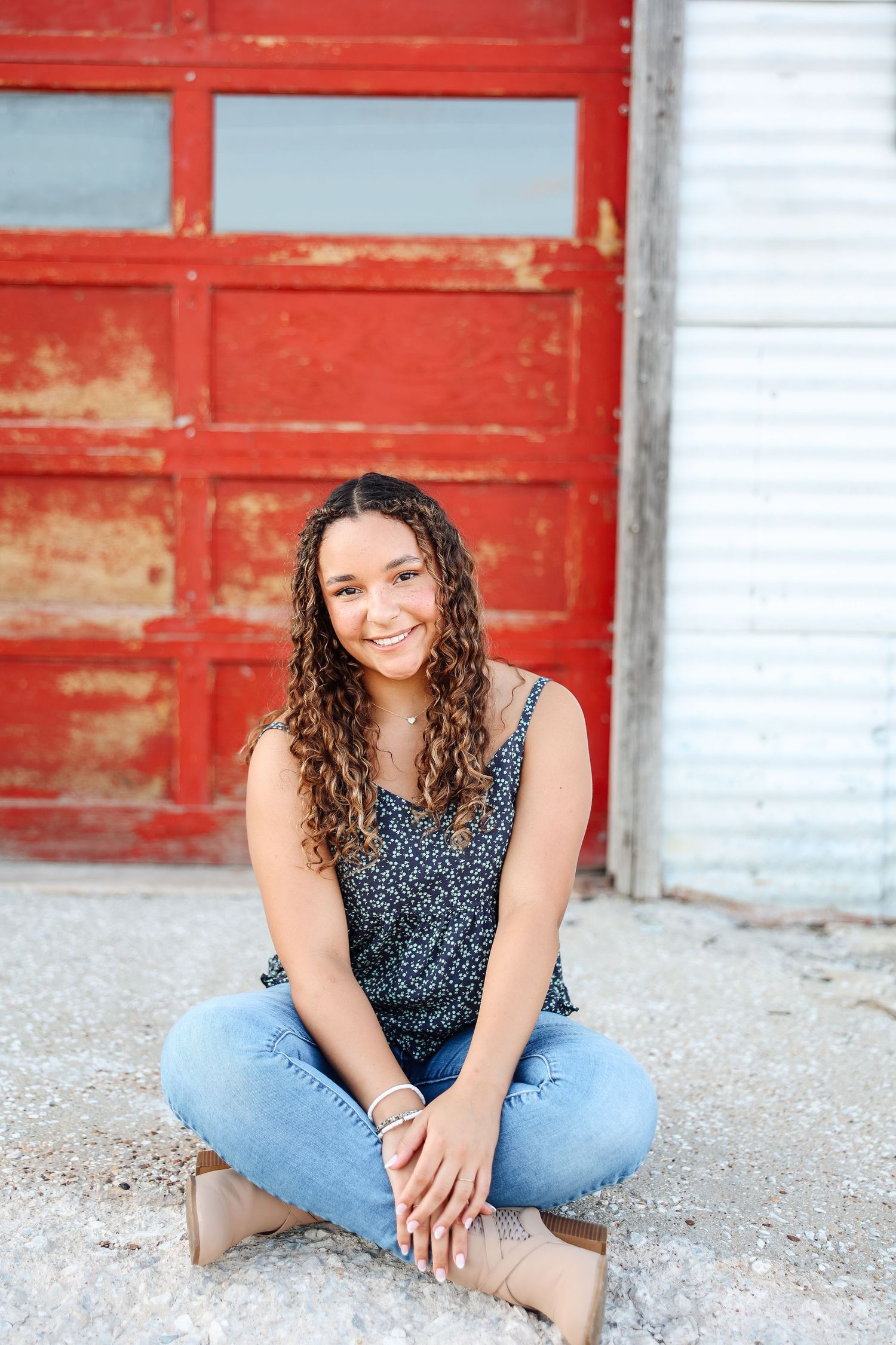 Young woman with curly hair sits cross-legged in front of a rustic red door wearing jeans and floral top.