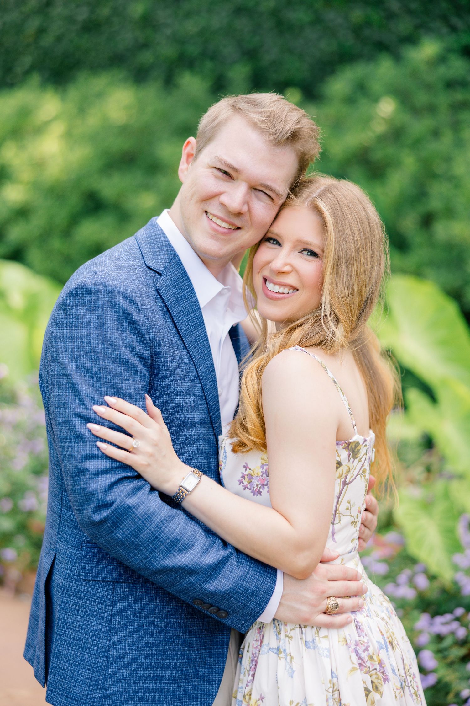 A smiling couple embraces outdoors, man in blue blazer and woman in floral dress, surrounded by greenery at Poetry Garden Dallas Arboretum