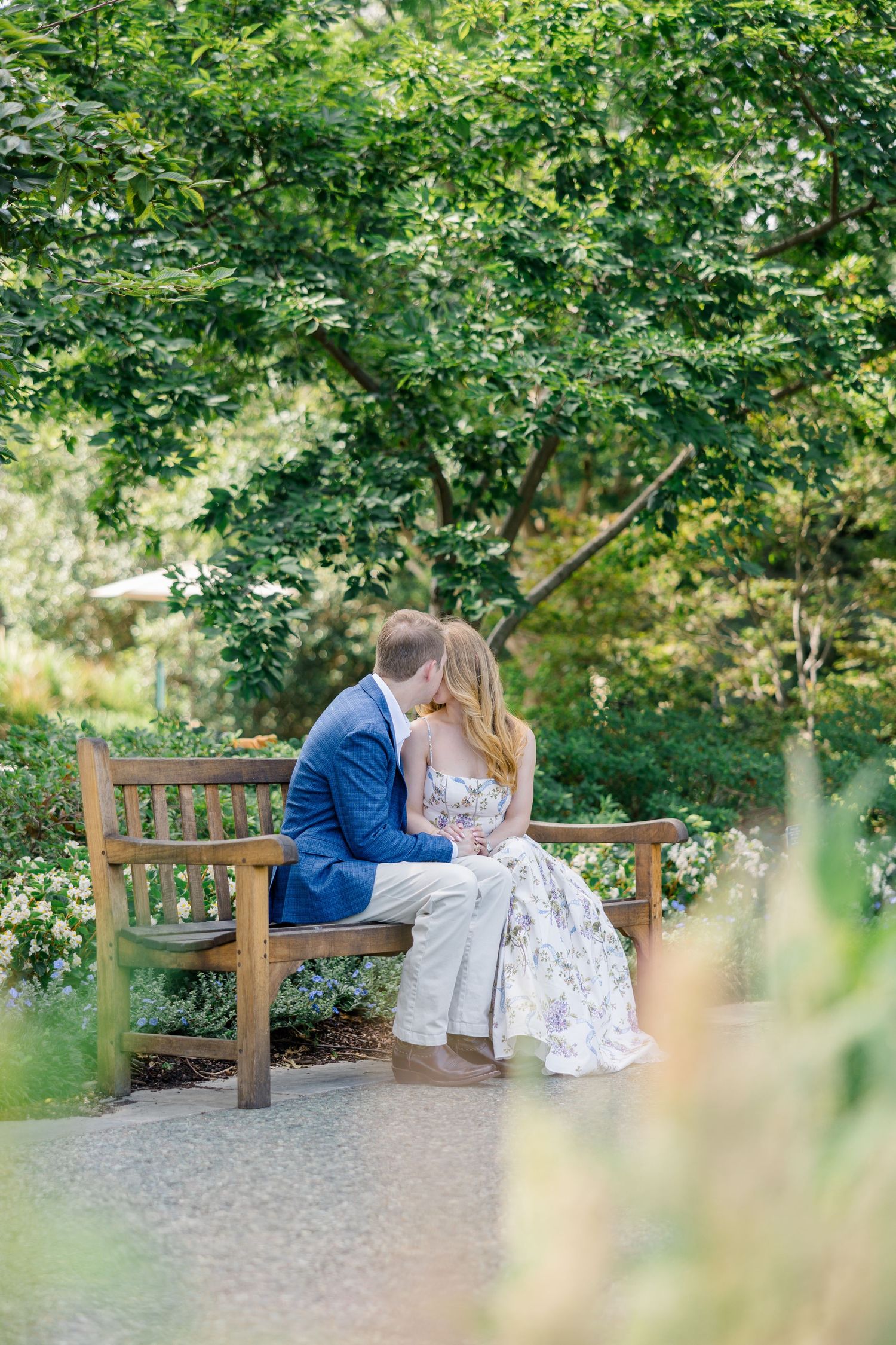 Couple sharing a romantic kiss on a wooden garden bench surrounded by lush green trees and blooming flowers at Dallas arboretum