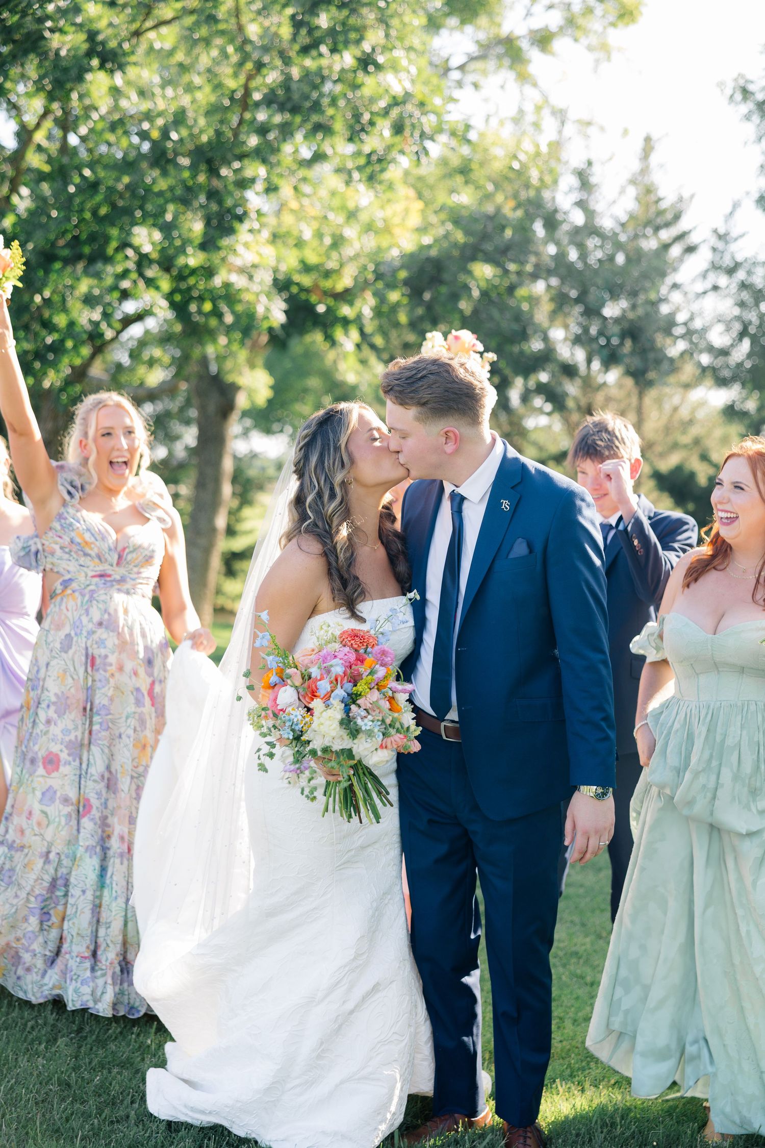 Wedding party in mint green floral bridesmaid dresses and navy suits celebrate joyfully outdoors on a sunny day. The Delanie Venue Forney TX