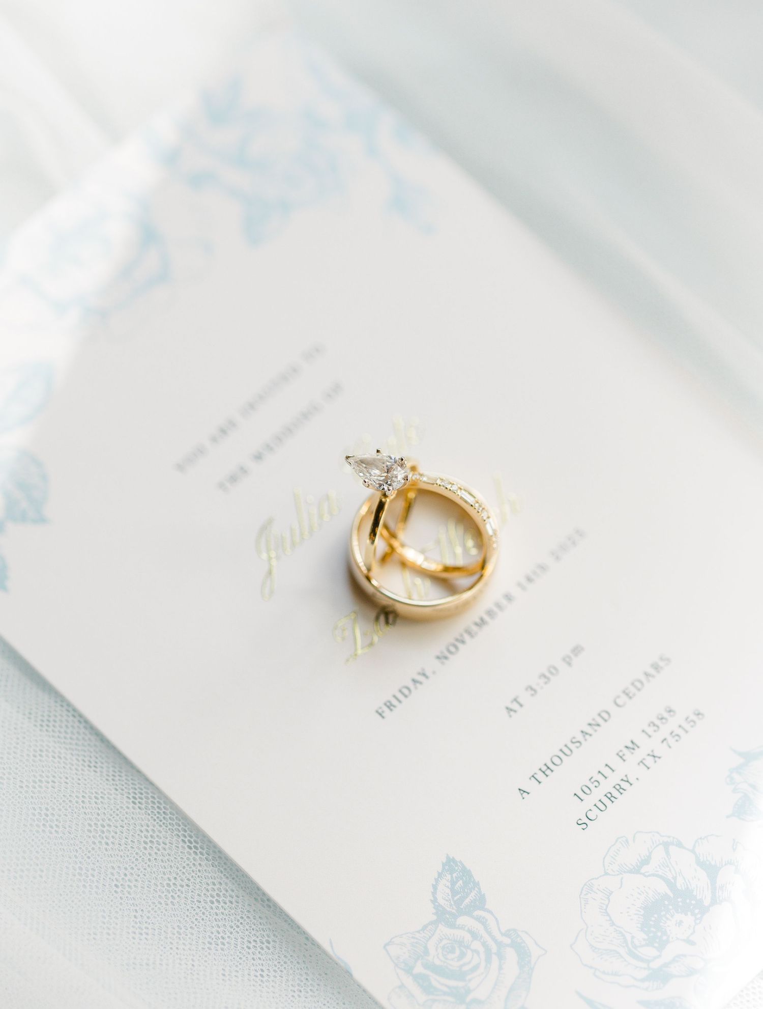 Two gold wedding rings rest elegantly on white paper with delicate blue floral designs. East Texas Wedding Photographer