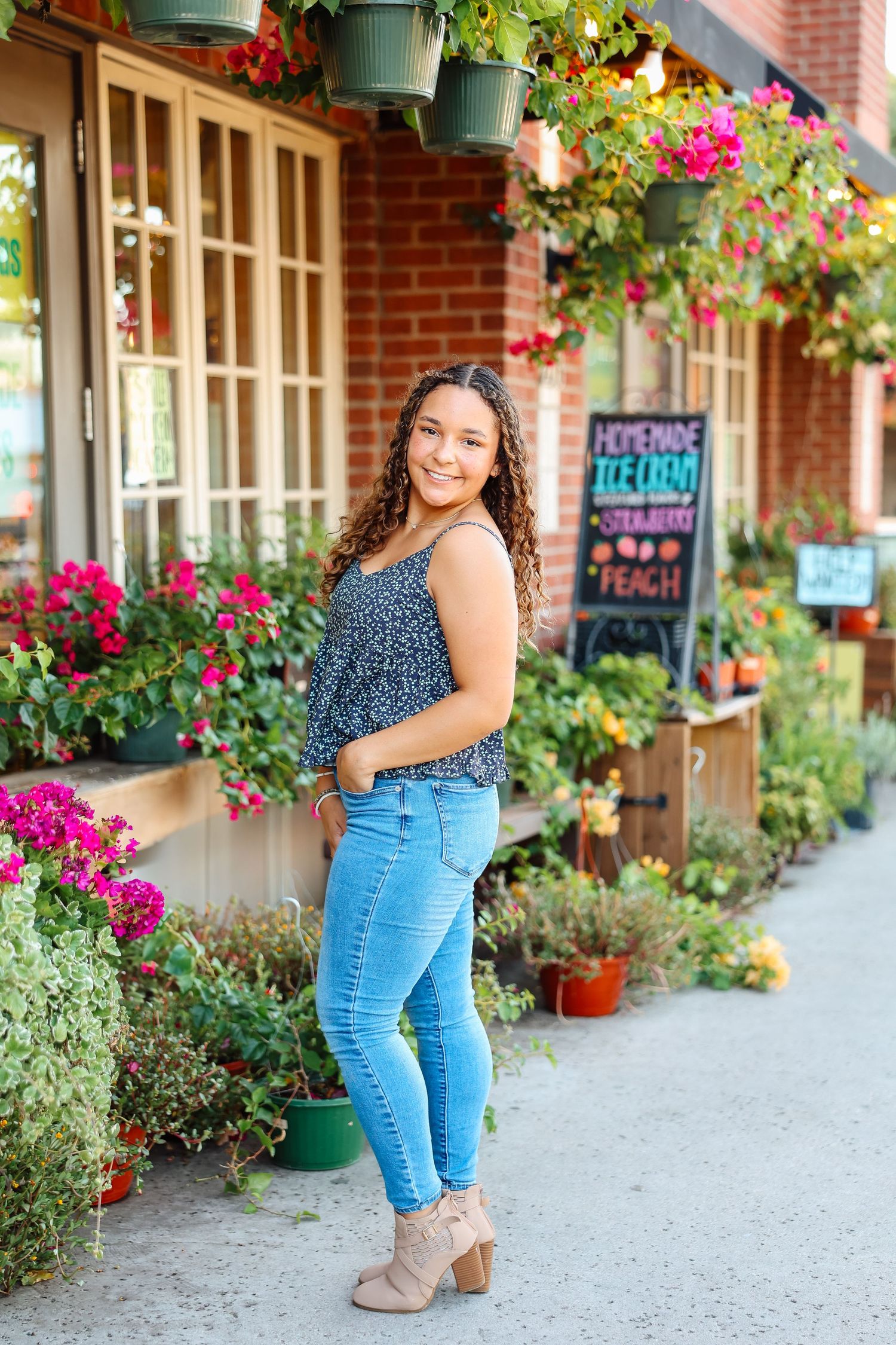 Person in polka dot top and jeans poses by brick storefront with bright pink flowers and greenery.