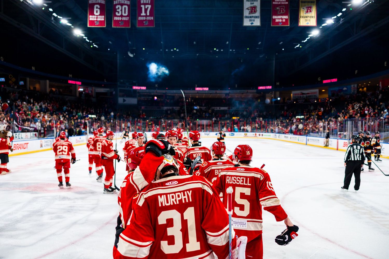 Hockey players in red jerseys celebrate on the ice during a game with championship banners visible in the arena rafters.