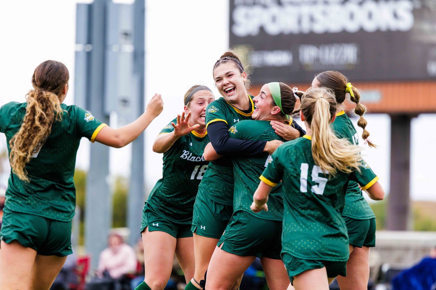 Women's rugby team celebrates together on the field in green uniforms after scoring during a match.