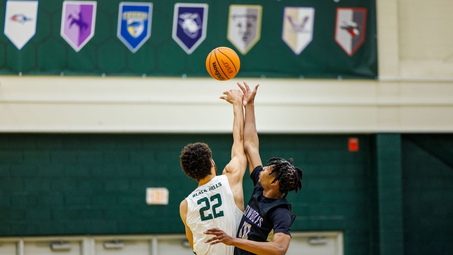 Basketball game action in a high school gymnasium with team banners displayed on the wall.