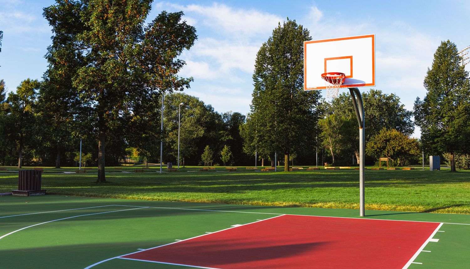 Outdoor basketball court with red paint and hoops set against a green park landscape.