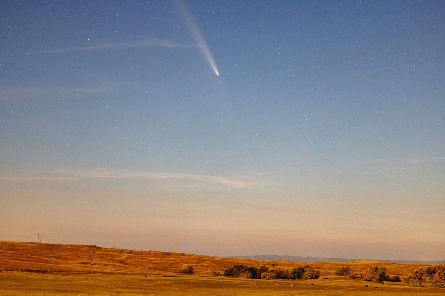 Comet NEOWISE streaks across golden sunset sky over rolling prairie landscape.