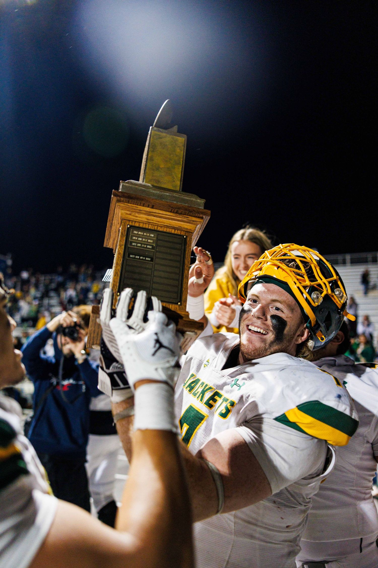 Football players in green and yellow uniforms celebrate with a trophy after winning a championship game.