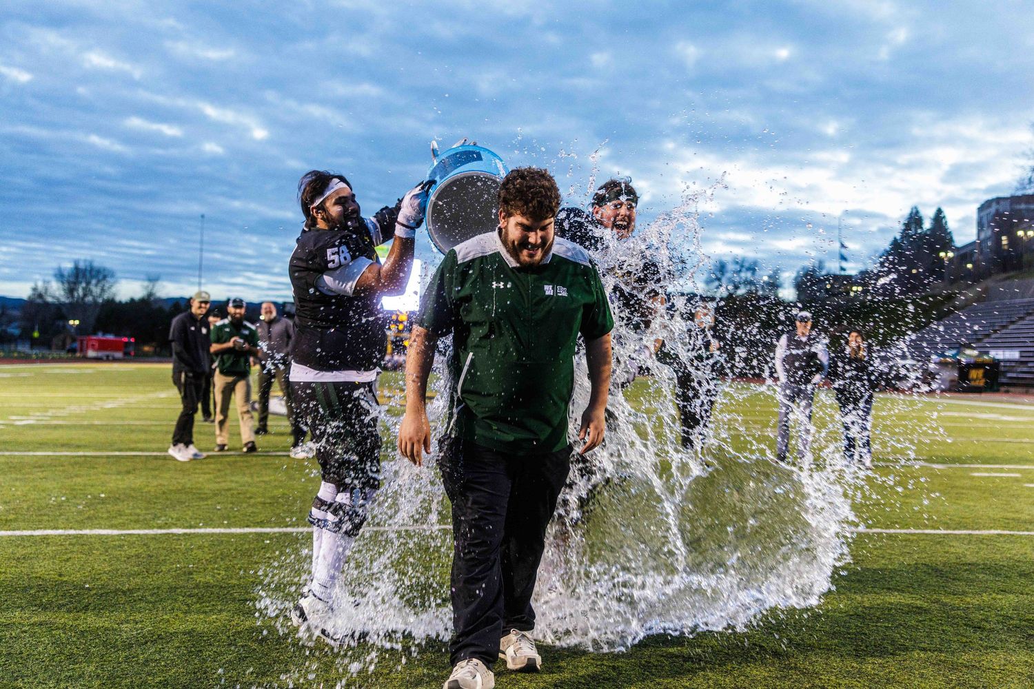Water splashes across a sunny field as people run through sprinklers on a grassy lawn.