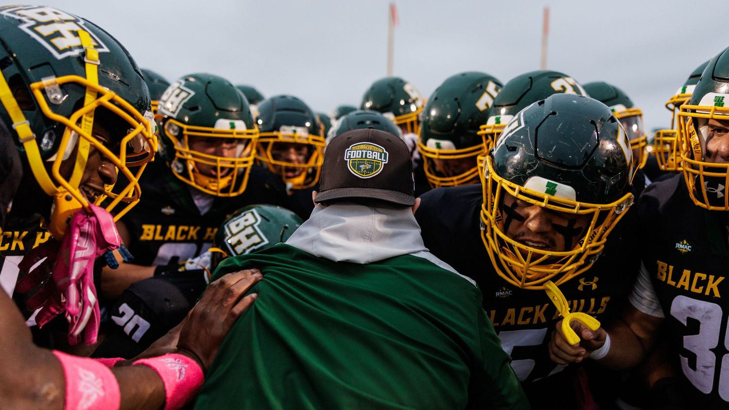 Football players in black and gold uniforms huddle together during a game while wearing pink accessories for breast cancer awareness.