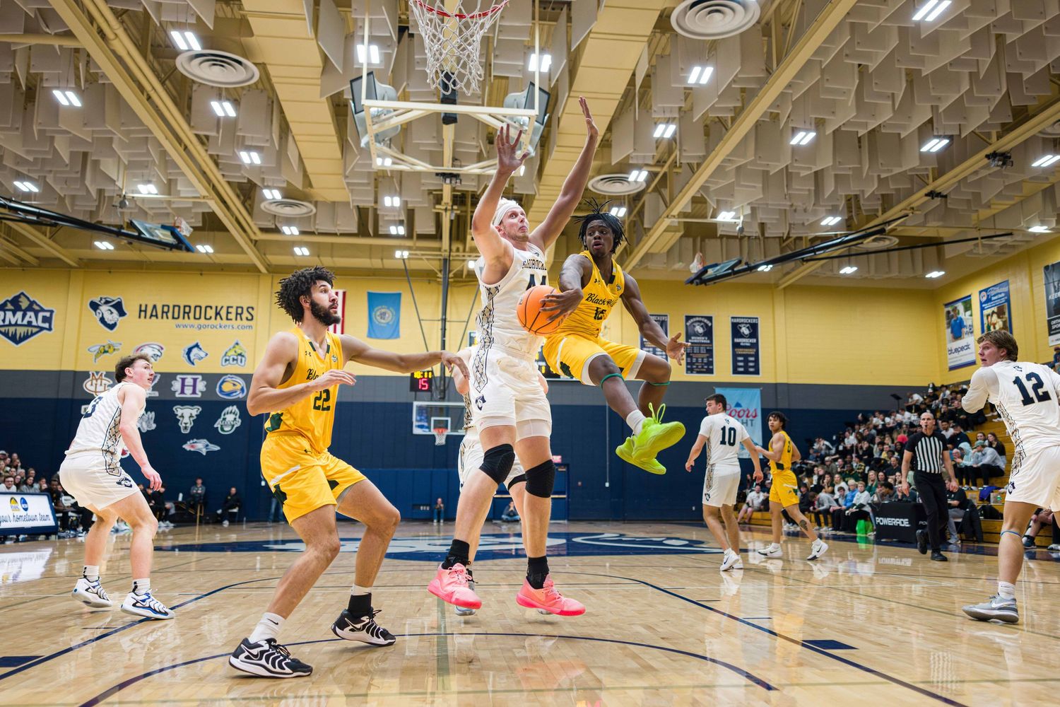 Basketball players compete for a rebound during an intense game on a yellow and blue court with spectators watching.