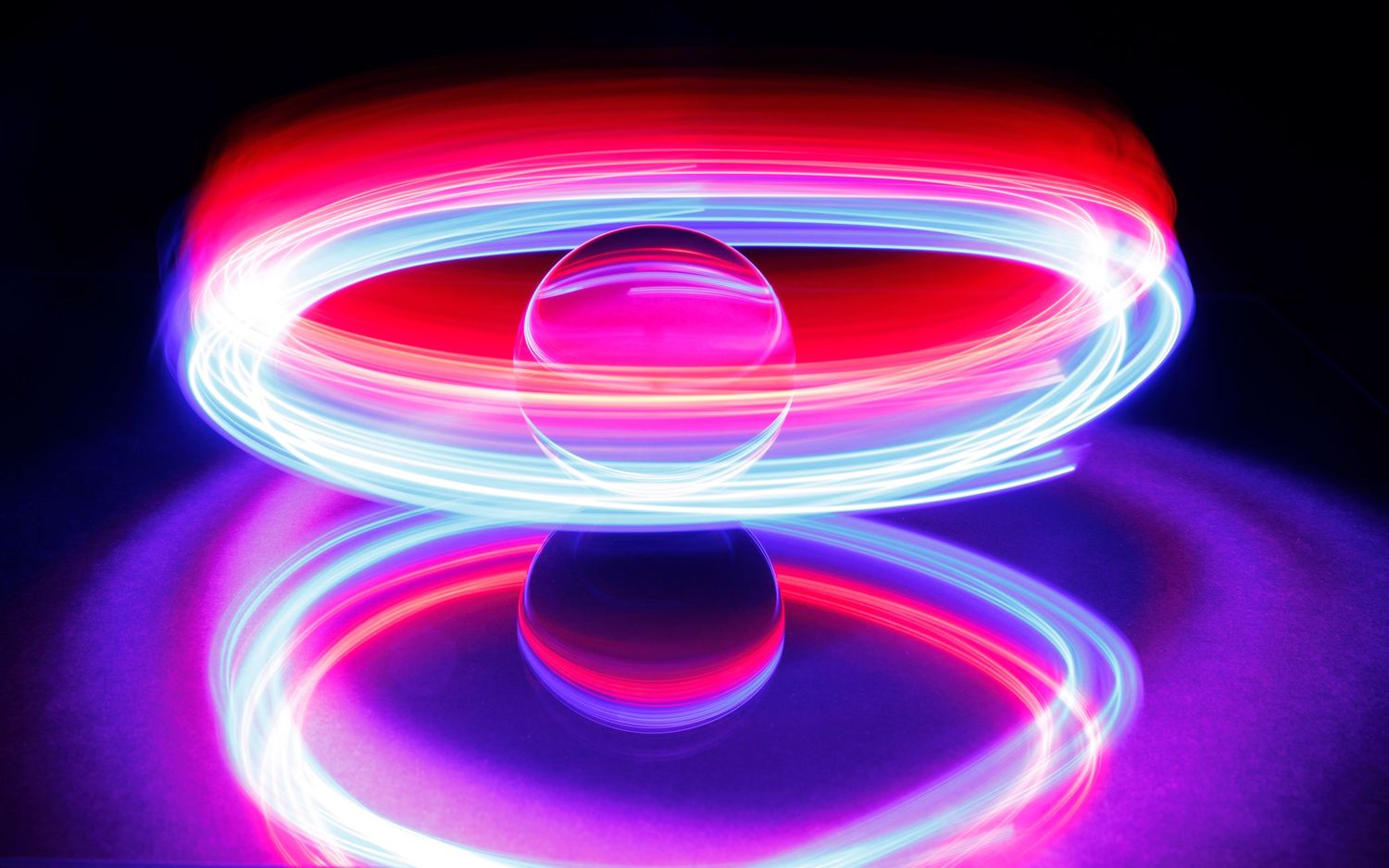Long exposure light painting creates glowing spiral rings in pink and blue against black background.