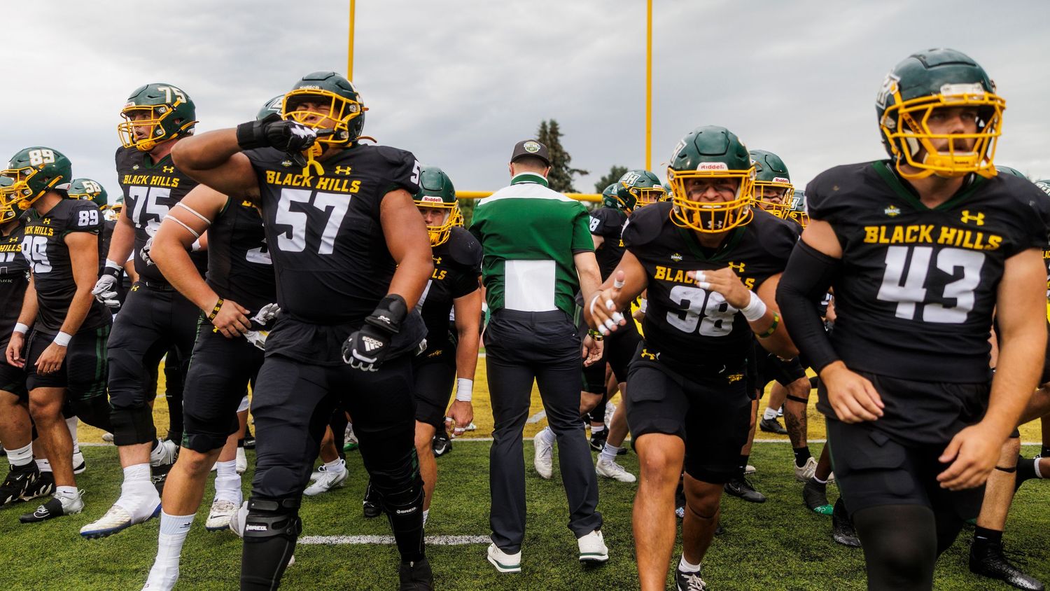 Football players in black uniforms and gold helmets lined up on the practice field.