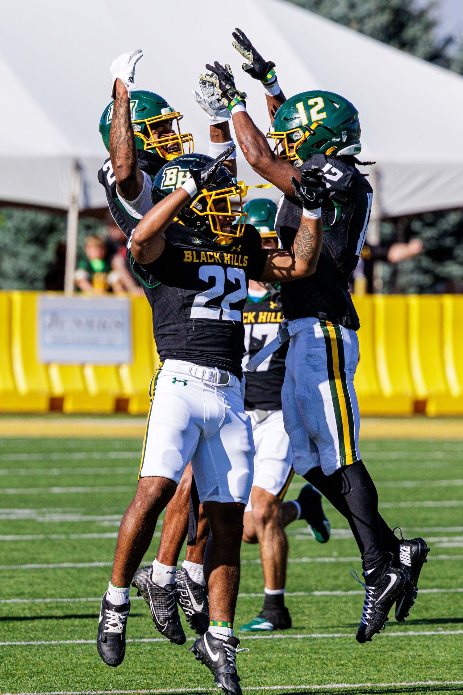Football players in black and gold uniforms practice defensive drills on a sunny field during training camp.
