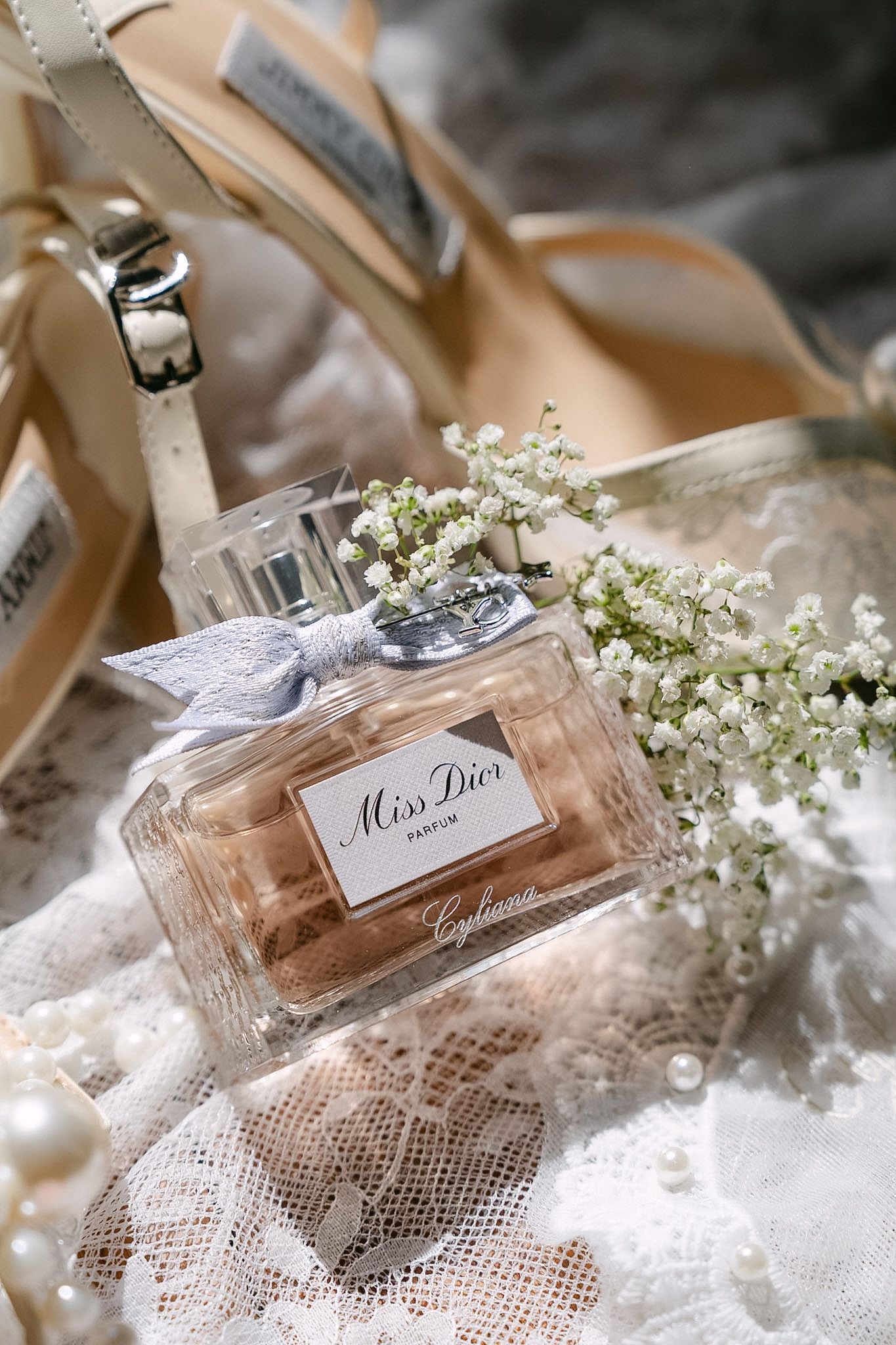 Miss Dior Parfum bottle with baby's breath flowers, pearl jewelry, and ivory heels on lace fabric.