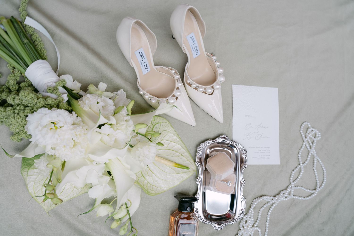 Elegant bridal flat lay with pearl-adorned heels, white floral bouquet, perfume, and wedding invitation on sage green fabric.